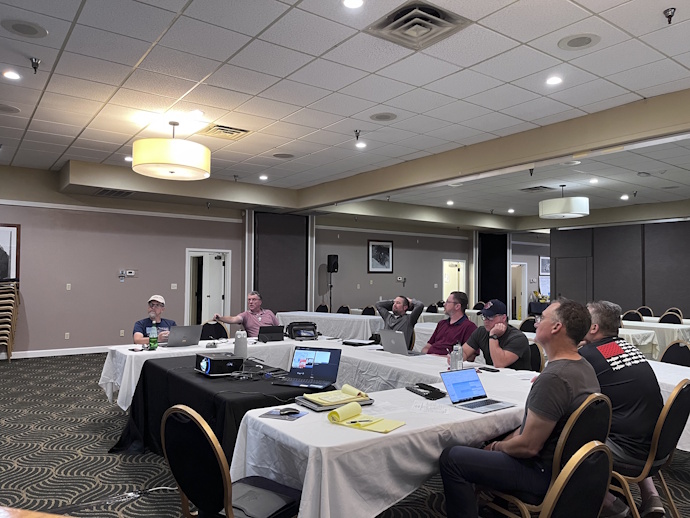Seven men at a meeting sitting at tables in a U shape configuration looking at one speaking or towards the front, a laptop and projector in the middle of the U.
