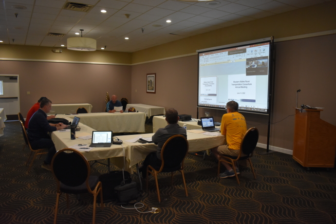 People meeting around tables facing projector screen virtual participants in upper right corner