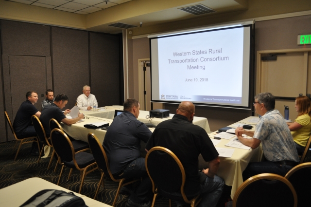Sean Campbell welcomes WSRTC Steering Committee members at the start of the 2018 annual meeting. A group of people seated at tables in a U shape with a projector screen in front, slide on screen with text: Western States Rural Transportation Consortium Meeting.