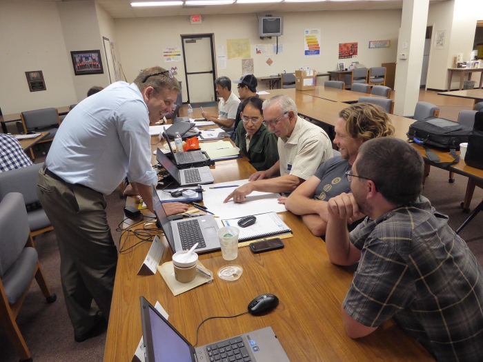 Students from Caltrans Districts 2 and 5 review their lab work with instructor Phil Isaak. (Left to Right: Phil Isaak, Steven Gee, Mike Beyer, Lonnie Hobbs, Keith Koeppen) An instructor stands in front of a table across from several seated students. The student in the middle is pointing to a paper on the table.