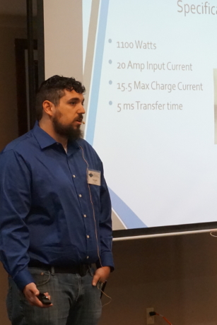 A man speaks beside a projector screen displaying 1100 Watts, 20 Amp Input Current, 15.5 Max Charge Current, 5 ms Transfer time