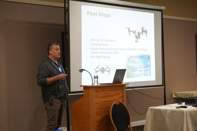 Tony Leingang A man speaks behind a podium next to a projector screen displaying a picture of a drone and the words First Steps