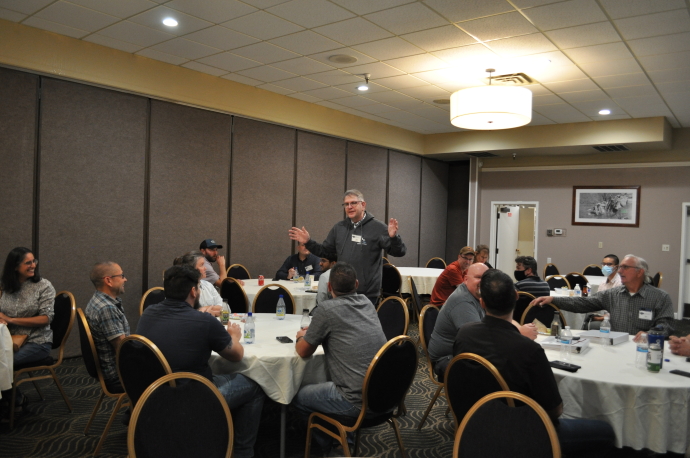 A man gesturing with arms open standing in the middle of a group of people seated at round tables.