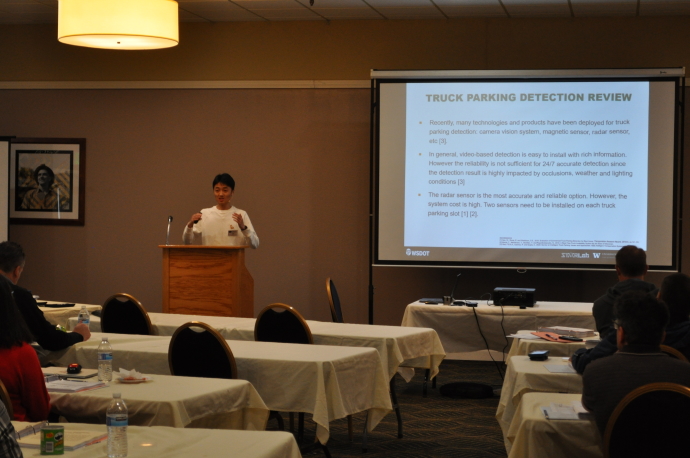Hao (Frank) Yang A speaker presents at a podium beside a screen showing Truck Parking Detection Review, audience watches