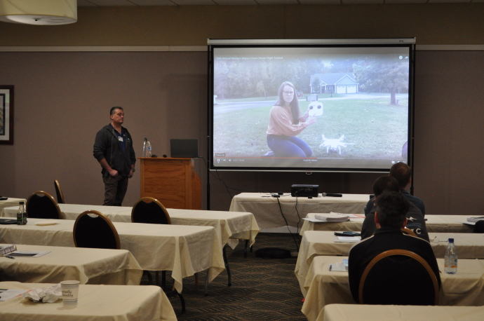 Alyssa Ryan A woman is shown in a video holding a drone, a man stands beside the projector screen, audience watches