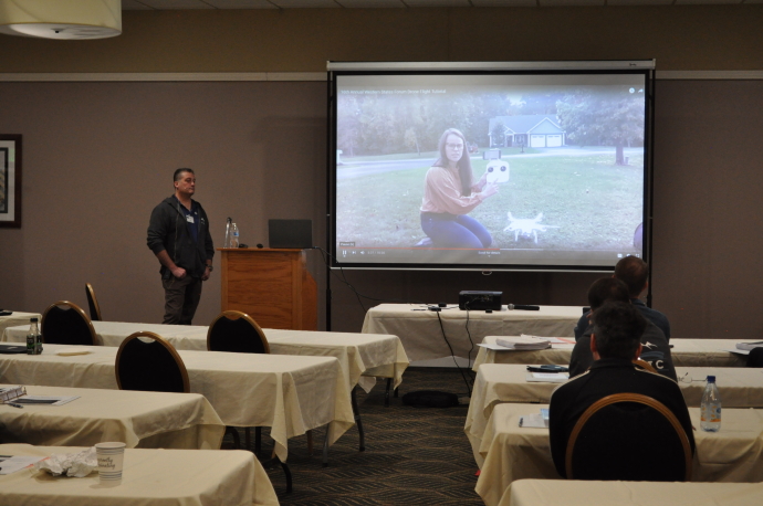 A woman is shown in a video holding a drone, a man stands beside the projector screen, audience watches