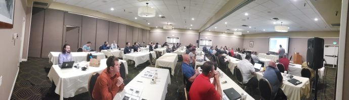 Group of people seated in rows at tables listening to speaker and watching a slide presentation.