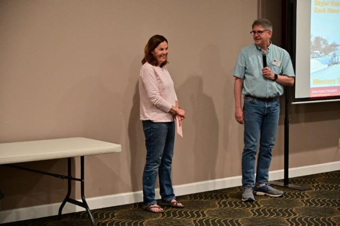 A woman (left) and a man (right) stand in front of a projector screen.