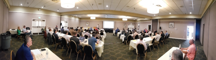 An audience seated at tables arranged classroom style watching a speaker in the front of the room by a screen.