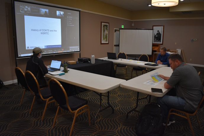2024 WSRTC Annual Meeting Three people seated at tables listen intently. A projector screen at the front shows online meeting participants and a slide with text: History of COATS and the WSRTC.