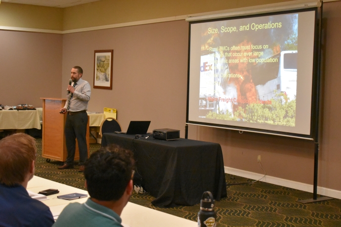 Jeremiah Pearce A man holds a microphone while standing beside a projector screen. Text on slide is Size, Scope, and Operations, image on slide is a burning semi-truck.
