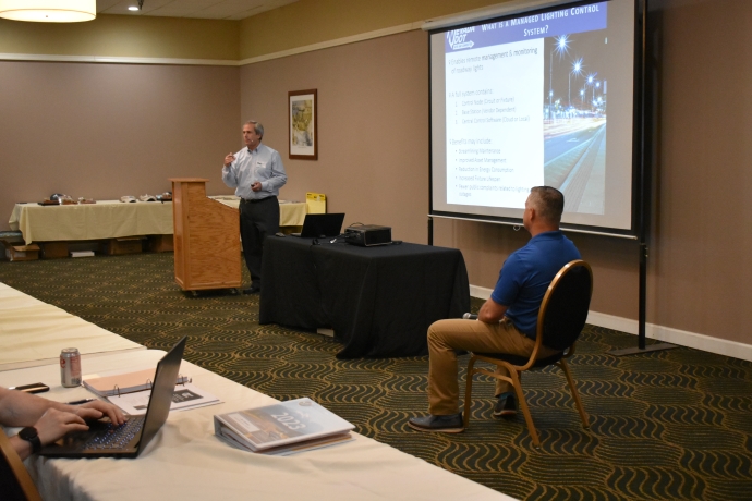 Eric MacGill (left) and Kevin Maxwell (right) A man presents to the left of a projector screen. Another man is seated across from him.