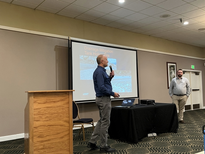 Jeff Williams (left) and Cody Oppermann (right) Two men stand on either side of a projector screen. The one on the left is speaking into a microphone.