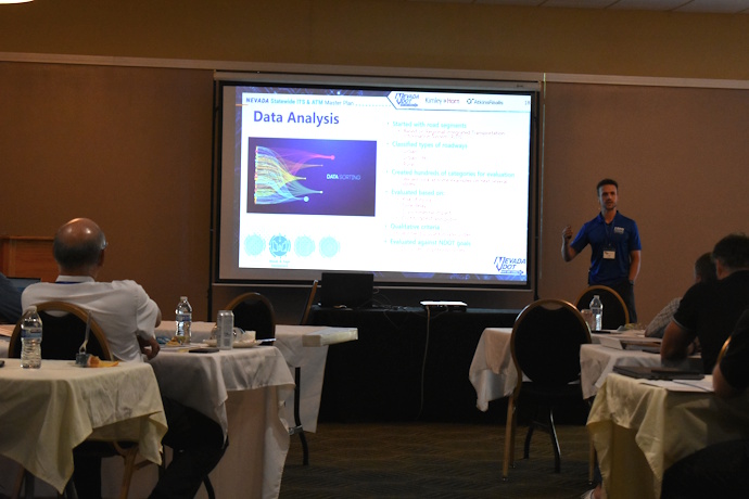A man points with his right hand while speaking beside a projector screen.
