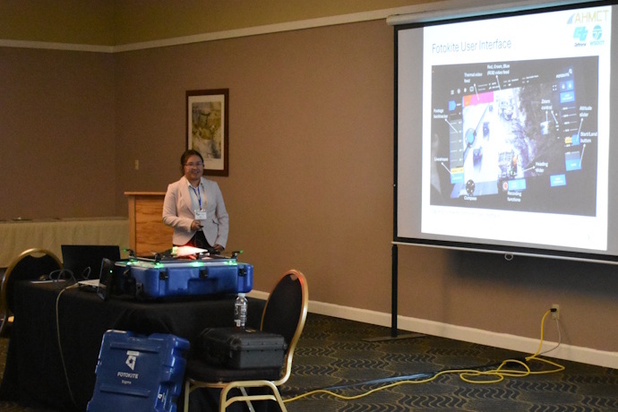 A woman with dark hair wearing a light pink blazer stands beside a projector screen with slide showing image and 'Fotokite User Interface'. In front of her on a table are three cases and a Fotokite drone.