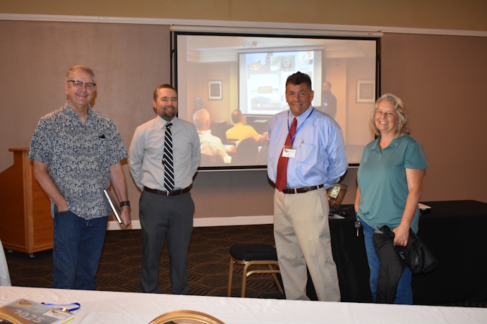 Three men and a woman stand smiling in front of a projector screen. The two men in the middle are wearing ties and dress clothes. The man on the left has a laptop in his hand and is wearing glasses.