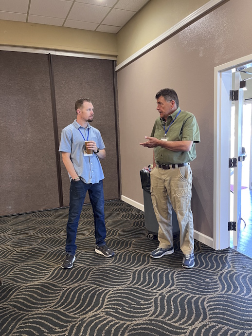 Two men stand talking to each other. The one on the right in a green shirt gestures to the other who is wearing a blue shirt and holding a paper coffee cup.