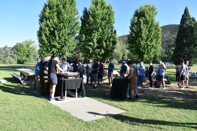 People outside lined up in a food buffet line. Large trees in the background, sun dappled grass.