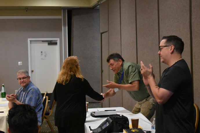 A woman with red hair shakes hands with a dark-haired man while handing him a box. A man standing to the side is clapping as is a seated man on the other side.
