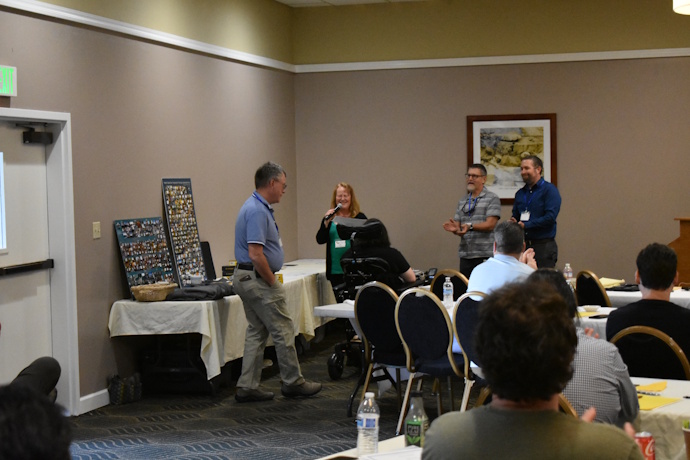 A man humbly walks toward a woman with a microphone and two men clapping.