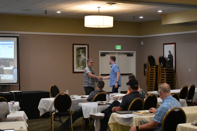 A man shakes hands with another man while handing him a box. An audience looks towards them.