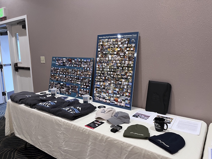 On a table: two large photo collages, Forum and WSRTC branded sweatshirts, mugs, coasters, ball caps, beanie hats, multi-tools, black padfolio.