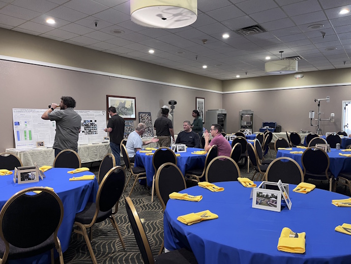 Round tables with framed photographs, yellow napkins with silverware tucked in, bright blue tablecloths, several people seated and milling around looking at the equipment, posters, and materials on tables against the wall.